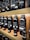 Multiple bottles of fragrance oil are neatly arranged on wooden shelves. The bottles vary in size and have labels with text both in English and another language. The lighting is warm, highlighting the sleek, dark-colored containers.