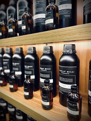 Multiple bottles of fragrance oil are neatly arranged on wooden shelves. The bottles vary in size and have labels with text both in English and another language. The lighting is warm, highlighting the sleek, dark-colored containers.