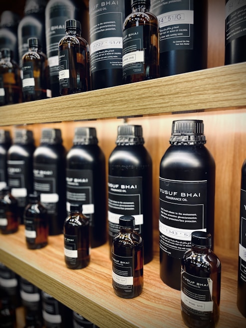 Multiple bottles of fragrance oil are neatly arranged on wooden shelves. The bottles vary in size and have labels with text both in English and another language. The lighting is warm, highlighting the sleek, dark-colored containers.