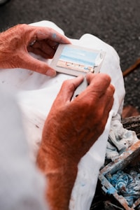 Hands of an elderly person painting a small picture of a beach scene on a miniature canvas, with paint-stained fingers holding a brush. The canvas is placed on a white piece of cloth, and a palette with blue paint is visible nearby.