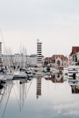 A tranquil marina featuring several moored sailboats and yachts with a distinctive black and white striped lighthouse in the background. The water is calm, creating reflective surfaces for both the vessels and the surrounding architecture.
