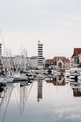 A tranquil marina featuring several moored sailboats and yachts with a distinctive black and white striped lighthouse in the background. The water is calm, creating reflective surfaces for both the vessels and the surrounding architecture.