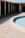 A drained pool with a light blue interior. Surrounding it is a circular concrete patio area. In the background, there is a structure with white support columns casting shadows. The area appears deserted and untouched.