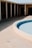 A drained pool with a light blue interior. Surrounding it is a circular concrete patio area. In the background, there is a structure with white support columns casting shadows. The area appears deserted and untouched.