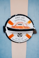 A circular emblem with a black outline and an orange and white color scheme features a boat at the center. The text 'AIDÉZ LES SAUVETEURS' is written on the top, and 'STÈ NLE DE SAUVETAGE EN MER' is inscribed at the bottom. The design includes an anchor and crossed oars. The background has a vertical beige stripe flanked by blue on either side.