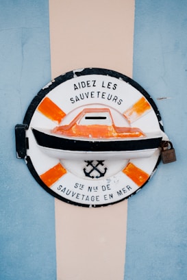 A circular emblem with a black outline and an orange and white color scheme features a boat at the center. The text 'AIDÉZ LES SAUVETEURS' is written on the top, and 'STÈ NLE DE SAUVETAGE EN MER' is inscribed at the bottom. The design includes an anchor and crossed oars. The background has a vertical beige stripe flanked by blue on either side.