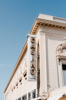 A vintage building features a vertical sign with the word 'CASINO' in large, lit letters, and 'CINEMA' below. The architecture includes ornate detailing near the roofline, and the facade has a cream color. The sky is clear and blue, enhancing the nostalgic feel of the setting.