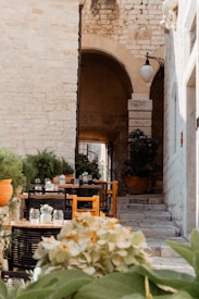 A cozy outdoor caf&eacute; setting with wooden tables and chairs is arranged along a narrow stone pathway. The tables are decorated with small vases containing flowers and glasses, giving a welcoming ambiance. Lush green plants and orange pots adorn the area. In the background, a stone archway and steps lead to a rustic building wall with an old-fashioned lamp hanging on it.