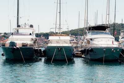 Three luxurious yachts are docked side by side in a marina, with clear blue water in the foreground. In the background, a row of small buildings and a green hillside can be seen under a clear sky.