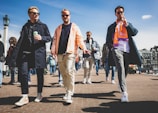 A group of casually dressed men walks along a sunny cobblestone street. The man on the left holds a can, wearing sunglasses and a dark coat. The man in the middle wears an orange jacket, while the man on the right sports an orange scarf and smoking a cigarette. People and historic buildings are visible in the background under a clear blue sky.
