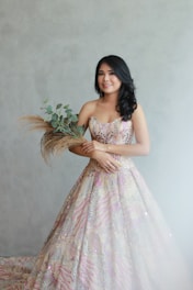 A young woman with long dark hair stands against a light gray background, wearing an intricately detailed, colorful gown with floral patterns. She holds a bouquet of greenery and dried grasses, smiling softly.