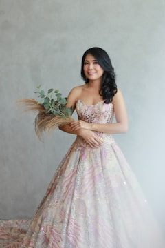 A young woman with long dark hair stands against a light gray background, wearing an intricately detailed, colorful gown with floral patterns. She holds a bouquet of greenery and dried grasses, smiling softly.