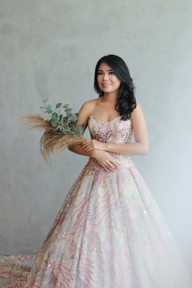 A young woman with long dark hair stands against a light gray background, wearing an intricately detailed, colorful gown with floral patterns. She holds a bouquet of greenery and dried grasses, smiling softly.