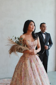 A woman in an elegant, intricately designed dress adorned with beads and floral patterns, holding a bouquet of dried flowers. A man in a formal black suit stands slightly behind her, both appearing to pose for a formal photo.