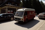 A red emergency vehicle, resembling an ambulance, is driving down a road in front of a building with stone and brick exterior. Alongside it are several parked cars. There is a forested area in the background, suggesting the setting is in a wooded or semi-rural area. The lighting indicates it is daytime with sunlight casting shadows on the vehicles and road.