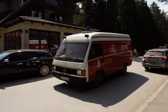 A red emergency vehicle, resembling an ambulance, is driving down a road in front of a building with stone and brick exterior. Alongside it are several parked cars. There is a forested area in the background, suggesting the setting is in a wooded or semi-rural area. The lighting indicates it is daytime with sunlight casting shadows on the vehicles and road.