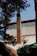A brick chimney extends from the roof of a building, surrounded by branches of a pine tree in the foreground. The building has a metal roof with a sloping design, and the sky is clear and blue.