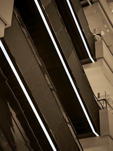 Several modern escalators are arranged in a parallel pattern within a sleek, contemporary interior space. The escalators have dark-colored sides with illuminated lines, accentuating the geometric design. The surrounding area features clean, angular architectural elements in light tones.