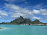 A serene tropical landscape featuring a turquoise lagoon with overwater bungalows situated beneath a lush, green mountain range. Fluffy white clouds float in a bright blue sky, creating a peaceful and idyllic scene.