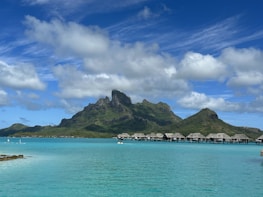 A serene tropical landscape featuring a turquoise lagoon with overwater bungalows situated beneath a lush, green mountain range. Fluffy white clouds float in a bright blue sky, creating a peaceful and idyllic scene.
