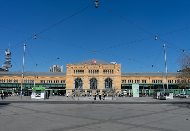 A large historic train station building with an intricate facade is situated under a clear blue sky. There are several people walking in the open square in front of the station, which is surrounded by modern buildings. Overhead, there are cables and streetlights for trams or electrical transportation.