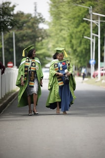 Two individuals wearing green graduation gowns and matching caps walk together on a paved road, with trees and street signs in the background. Both are adorned with colorful stoles featuring designs, indicating an academic achievement.