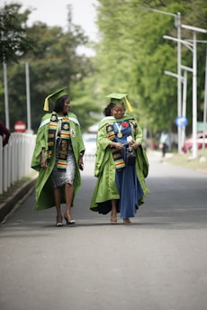 Two individuals wearing green graduation gowns and matching caps walk together on a paved road, with trees and street signs in the background. Both are adorned with colorful stoles featuring designs, indicating an academic achievement.