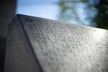 An engraved stone memorial with names, dates, and locations etched into it, including references to Auschwitz and Sobibor. A small Star of David is visible at the top, indicating a Jewish memorial. The background is blurred, showing green foliage.