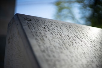 An engraved stone memorial with names, dates, and locations etched into it, including references to Auschwitz and Sobibor. A small Star of David is visible at the top, indicating a Jewish memorial. The background is blurred, showing green foliage.