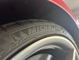 A close-up of a car tire with the 'Michelin' branding prominently displayed along with a small logo. The surrounding element of the car, possibly a wheel arch, is visible, and the tire appears new with clean tread patterns.