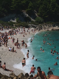 A crowded beach scene with numerous people sunbathing on the shore and swimming in the turquoise sea. The shoreline is pebbly, surrounded by lush green trees and rocky cliffs. Some people are standing in the waves while others are lying on beach towels.