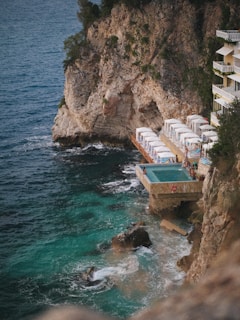 A rocky coastline features a small cliffside resort with a pool overlooking the ocean. White loungers with umbrellas line a wooden deck adjacent to the pool, inviting relaxation. The turquoise water crashes gently against the rocks, creating an idyllic and peaceful seaside retreat.