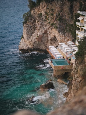 A rocky coastline features a small cliffside resort with a pool overlooking the ocean. White loungers with umbrellas line a wooden deck adjacent to the pool, inviting relaxation. The turquoise water crashes gently against the rocks, creating an idyllic and peaceful seaside retreat.