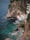 A rocky coastline features a small cliffside resort with a pool overlooking the ocean. White loungers with umbrellas line a wooden deck adjacent to the pool, inviting relaxation. The turquoise water crashes gently against the rocks, creating an idyllic and peaceful seaside retreat.