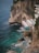 A rocky coastline features a small cliffside resort with a pool overlooking the ocean. White loungers with umbrellas line a wooden deck adjacent to the pool, inviting relaxation. The turquoise water crashes gently against the rocks, creating an idyllic and peaceful seaside retreat.