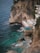 A rocky coastline features a small cliffside resort with a pool overlooking the ocean. White loungers with umbrellas line a wooden deck adjacent to the pool, inviting relaxation. The turquoise water crashes gently against the rocks, creating an idyllic and peaceful seaside retreat.