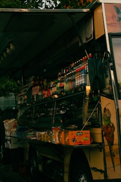 A dimly lit food truck with a variety of packaged goods and beverages displayed inside. The truck is adorned with colorful images of ice cream cones on the exterior, and it is parked on a street, partially obscured by dark shadows and trees.