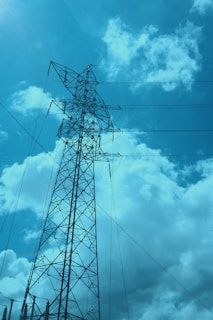 A tall metal electricity transmission tower rises against a backdrop of fluffy white clouds and a clear blue sky. Power lines extend outward from the tower, spanning across the scene.