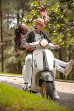 A man wearing a hat and a leather jacket is riding a white scooter on a tree-lined path. A woman sits behind him, smiling brightly. Both appear joyful and carefree, with the man holding a bouquet of flowers decorated with ribbons. They are surrounded by vibrant green foliage.