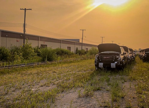 A row of dark-colored trucks with open hoods is parked on a grassy, unpaved area adjacent to a long industrial building. The sky is hazy with the sun setting or rising in the background, casting a warm golden glow over the scene. There are power lines and telephone poles running parallel to the building.