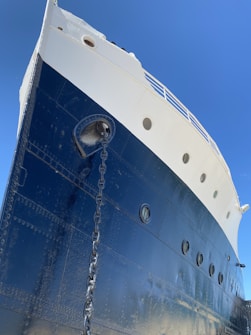 A large, imposing ship hull viewed from a low angle, emphasizing its size and the deep blue color of its metal surface. The ship has a sturdy anchor chain visible, and the hull features multiple small, circular windows. The upper portion is white, and a clear blue sky provides a contrasting background.
