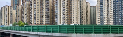 A collection of high-rise residential buildings with numerous windows and balconies. In the foreground, a green barrier runs along the top of an overpass, obstructing the lower portion of the buildings. The scene suggests an urban environment with dense housing.