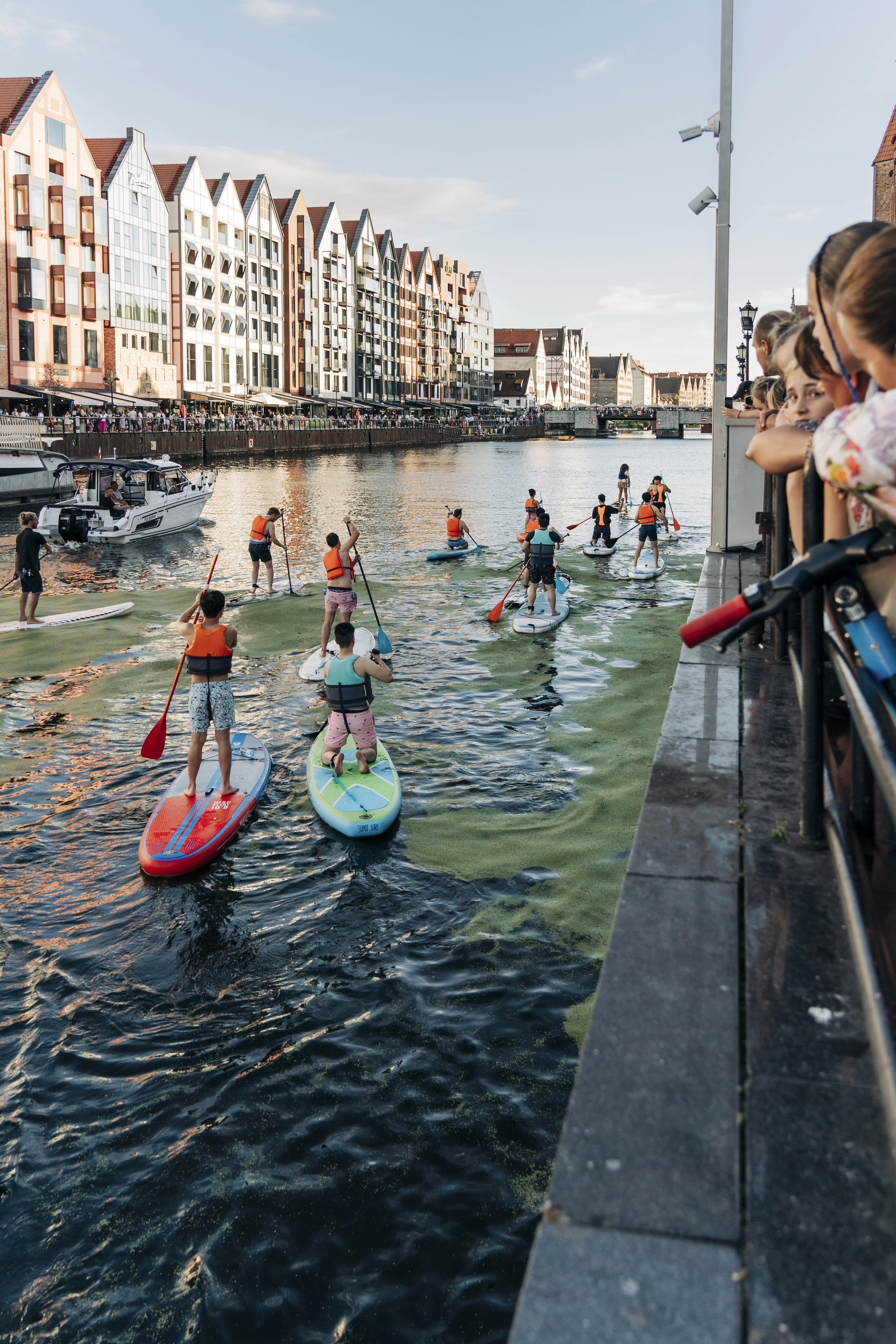 Children on paddleboards glide down Gdańsk's canal in front of the colorful gabled townhouses of the Long Embankment.