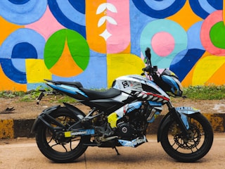 A black and blue sports motorcycle is parked on a pavement next to a brightly painted mural wall. The mural features a variety of geometric shapes in vibrant colors including blue, yellow, pink, and green. The motorcycle has decals featuring various patterns and symbols.