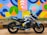 A black and blue sports motorcycle is parked on a pavement next to a brightly painted mural wall. The mural features a variety of geometric shapes in vibrant colors including blue, yellow, pink, and green. The motorcycle has decals featuring various patterns and symbols.