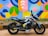 A black and blue sports motorcycle is parked on a pavement next to a brightly painted mural wall. The mural features a variety of geometric shapes in vibrant colors including blue, yellow, pink, and green. The motorcycle has decals featuring various patterns and symbols.