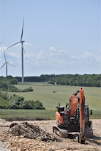 A construction site featuring an orange excavator parked on rocky ground in the foreground. In the background, several wind turbines are set across a vast, green landscape, bordered by trees under a clear blue sky.