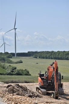 A construction site featuring an orange excavator parked on rocky ground in the foreground. In the background, several wind turbines are set across a vast, green landscape, bordered by trees under a clear blue sky.