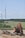 A construction site featuring an orange excavator parked on rocky ground in the foreground. In the background, several wind turbines are set across a vast, green landscape, bordered by trees under a clear blue sky.