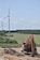 A construction site featuring an orange excavator parked on rocky ground in the foreground. In the background, several wind turbines are set across a vast, green landscape, bordered by trees under a clear blue sky.
